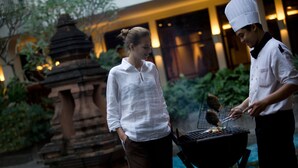 A person grilling outdoors near a pool with a temple-like structure in the background.
