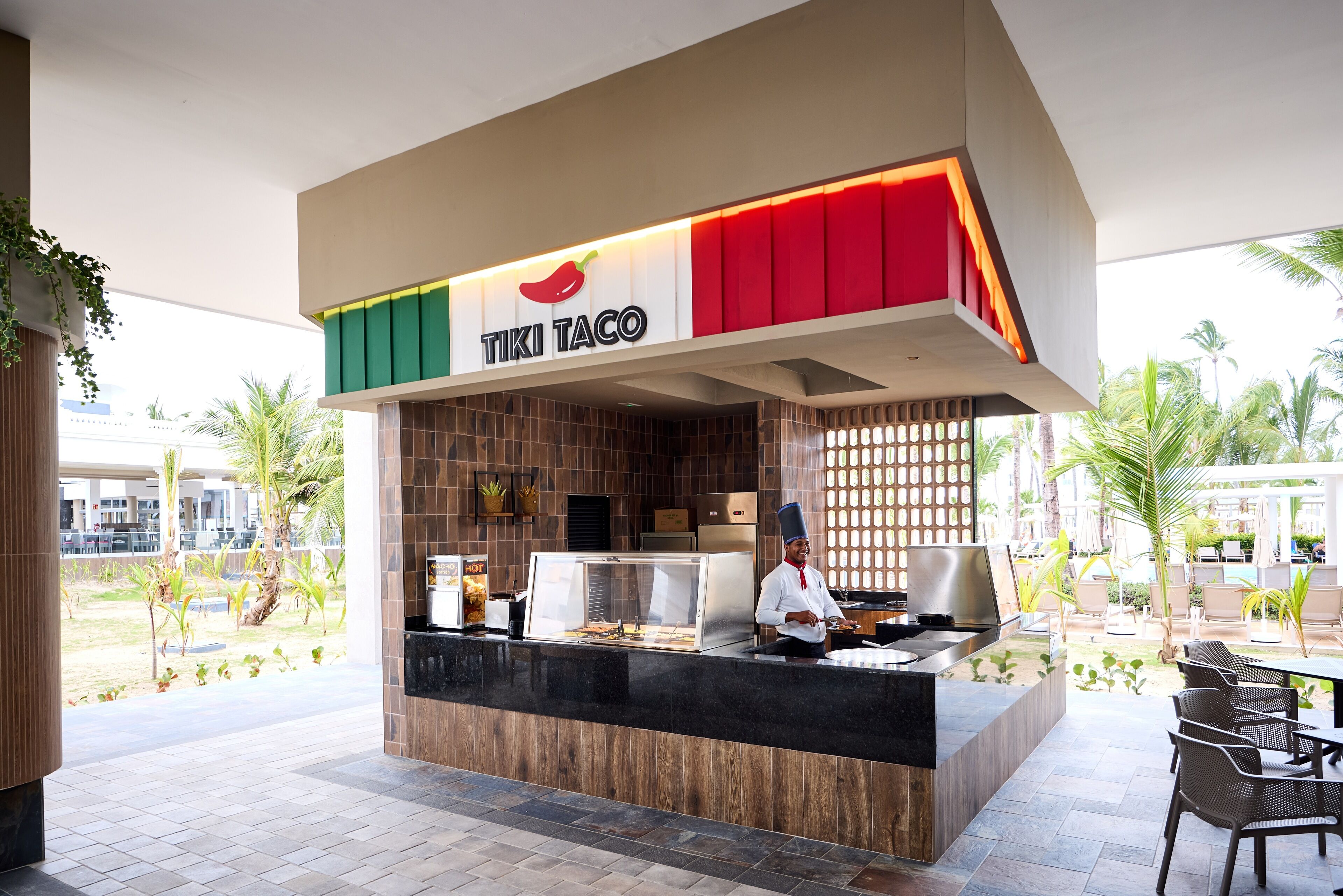 A TIKI TACO food counter with a chef, outdoor seating, and a view of palm trees.