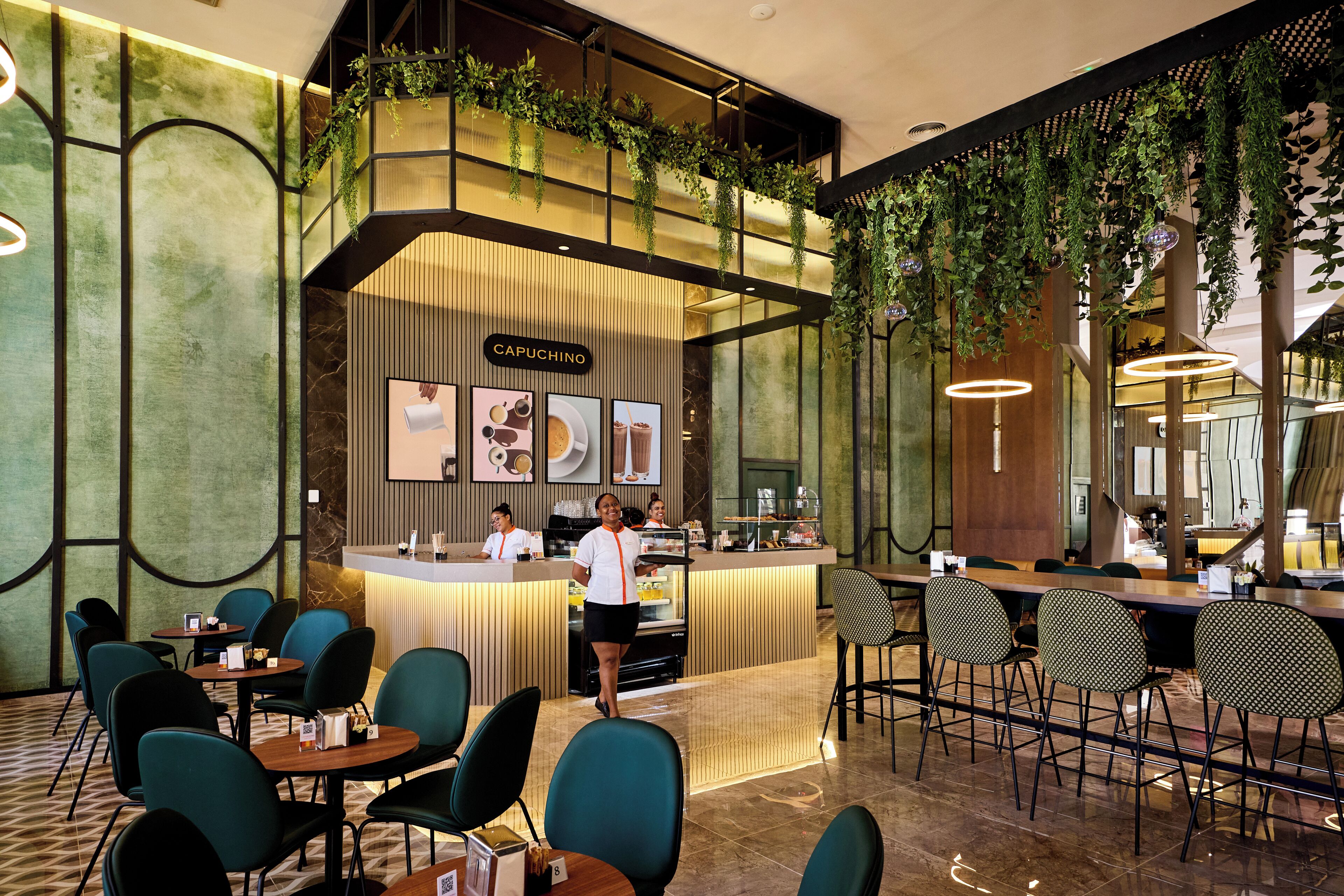 A modern cafe with a counter labeled "CAPUCHINO," featuring a variety of seating options and a decorative ceiling with hanging plants.