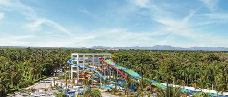 A water park with multiple slides and a pool area, surrounded by palm trees and mountains in the background.