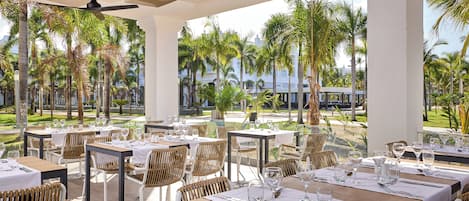 A restaurant with outdoor seating, white tables, wicker chairs, and a view of palm trees and buildings.