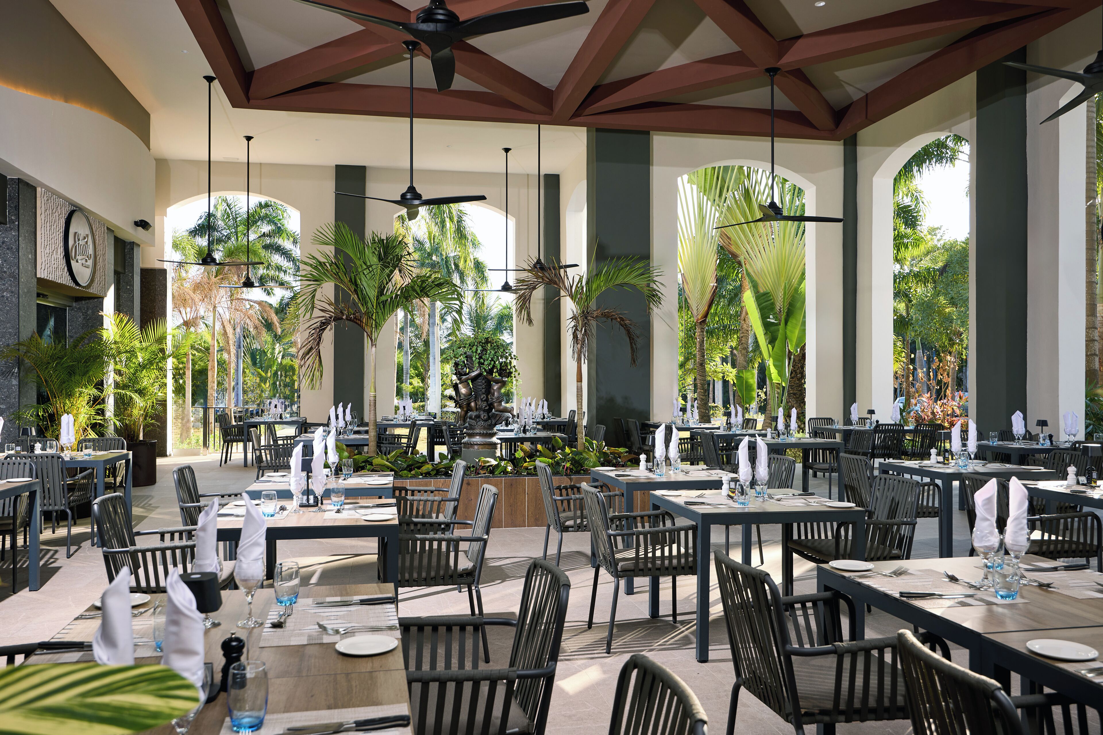 An outdoor dining area with multiple tables and chairs, palm trees, and a ceiling fan.