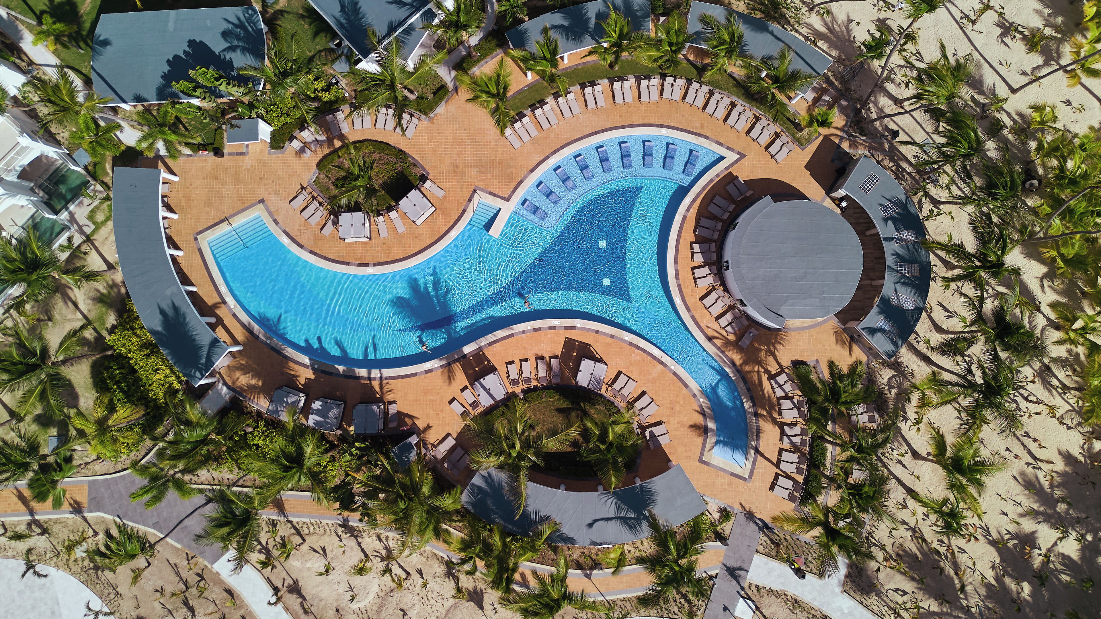 An aerial view of a resort with a large, curved swimming pool surrounded by palm trees and lounge chairs.
