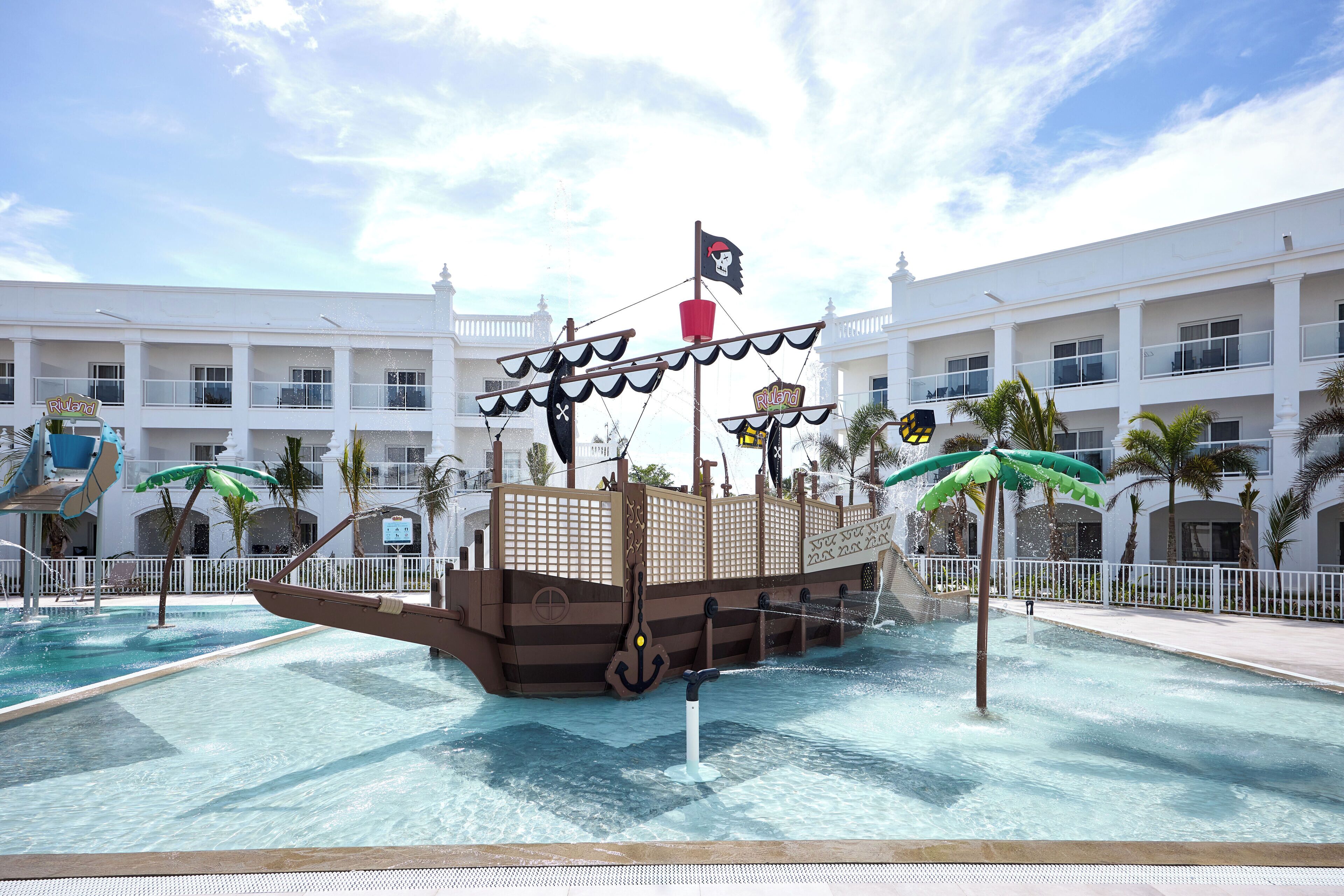 A pirate-themed water attraction with a ship structure in a pool, surrounded by a white building with balconies and palm trees.