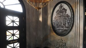 Una sala elegante con una gran lámpara de araña, un cuadro histórico enmarco y un diseño de suelo llamativo.