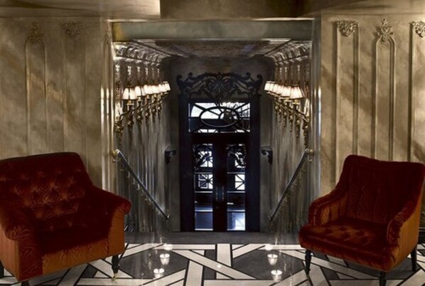 A grand entrance with a black and white geometric floor pattern, two red velvet armchairs, and a staircase with ornate railings and wall sconces.
