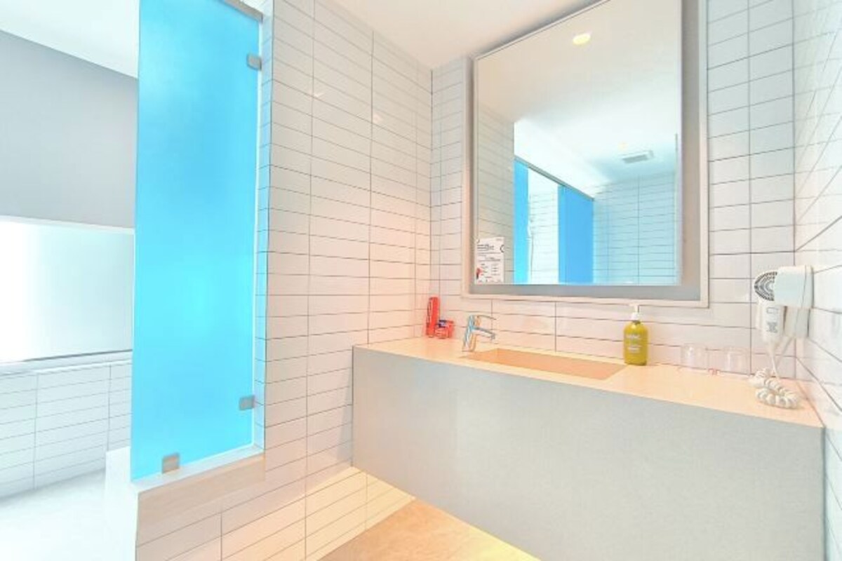 A modern bathroom with white tiled walls, a blue accent wall, a large mirror, a sink with a faucet, and a yellow bottle of soap.