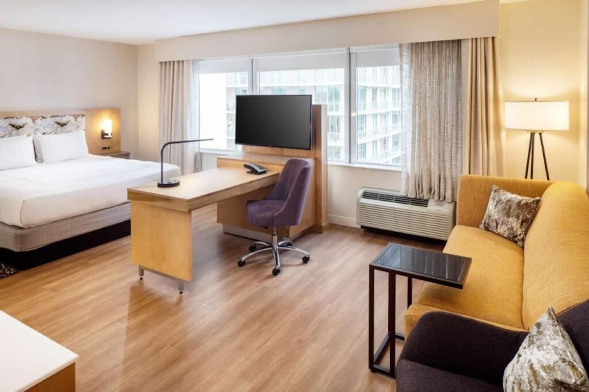 A hotel room with a bed, desk, chair, sofa, TV, and windows with curtains.