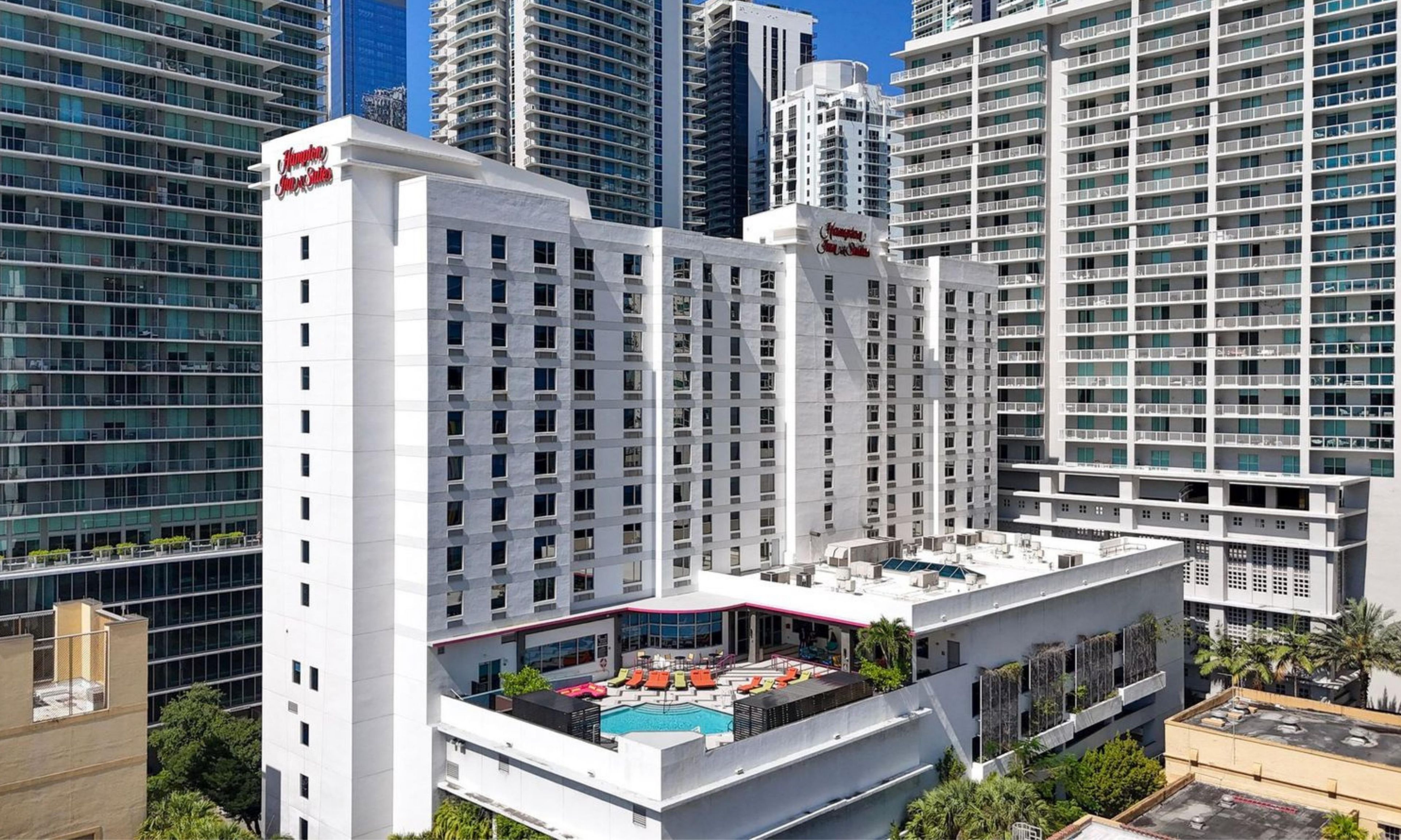 Un hotel di grandi dimensioni con numerosi piani, un'area piscina sul tetto e un cartello ben visibile con la scritta 'Hampton Inn & Suites'.