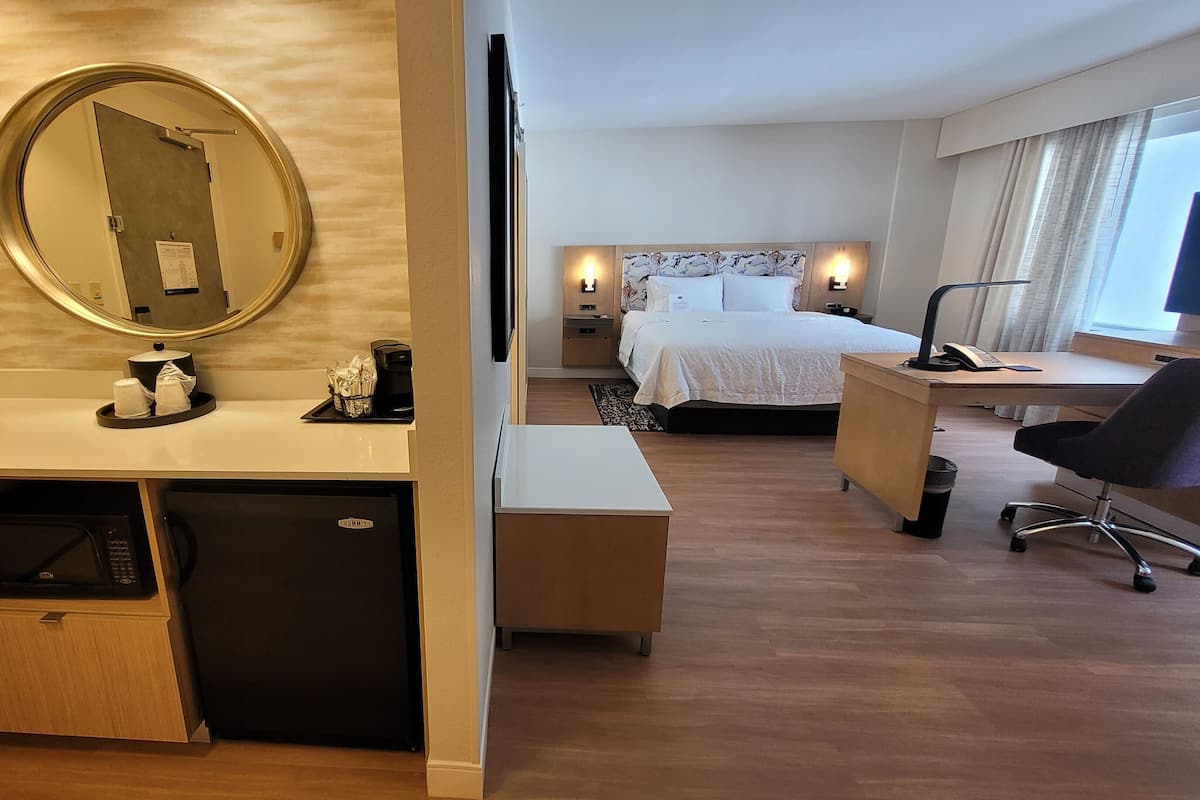 A hotel room with a wooden desk, a bed with white bedding, a large mirror, and a small sink with a coffee machine.