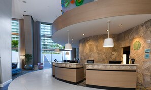 A hotel reception area with two reception desks, a large window with a view of buildings, and a stone wall.