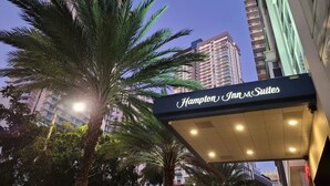 Een hotelschilt met de naam 'Hampton Inn & Suites' voor een achtergrond van hoge gebouwen en palmbomen.