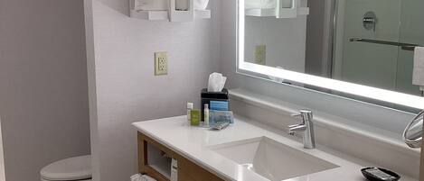 A modern bathroom with a large mirror, two sinks, and a toilet. There are towels hanging on the wall and a shelf with toiletries.