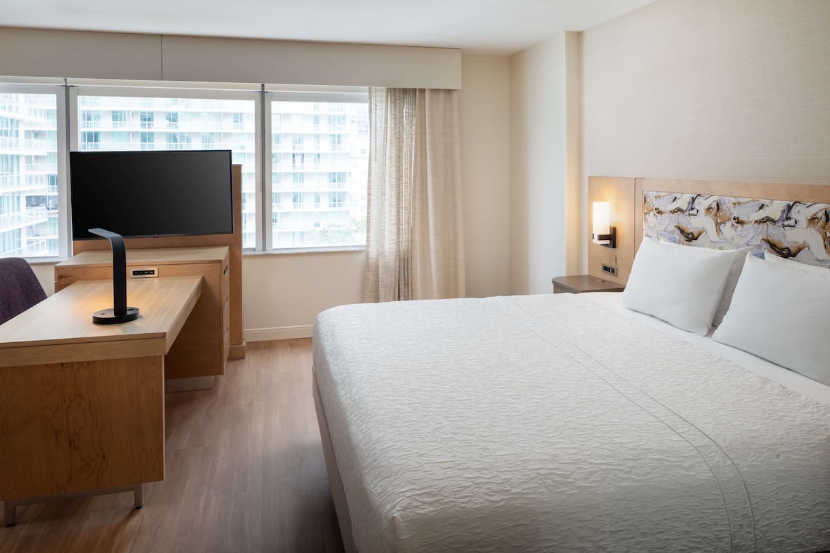 A hotel room with a bed, a desk with a flat-screen TV, a nightstand with a lamp, and a window with curtains.