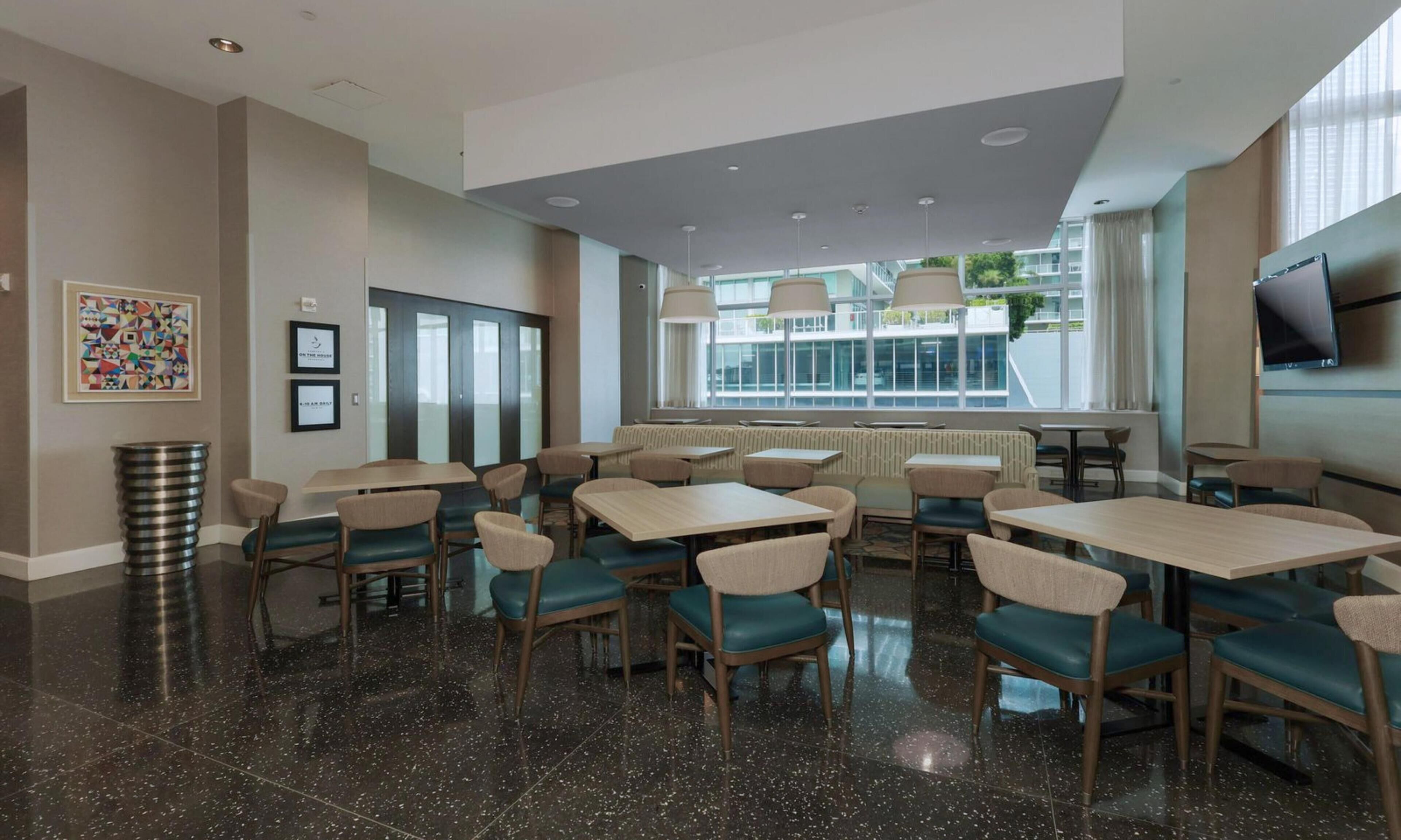 A modern dining area with multiple tables and chairs, a large window with a view, and a television mounted on the wall.
