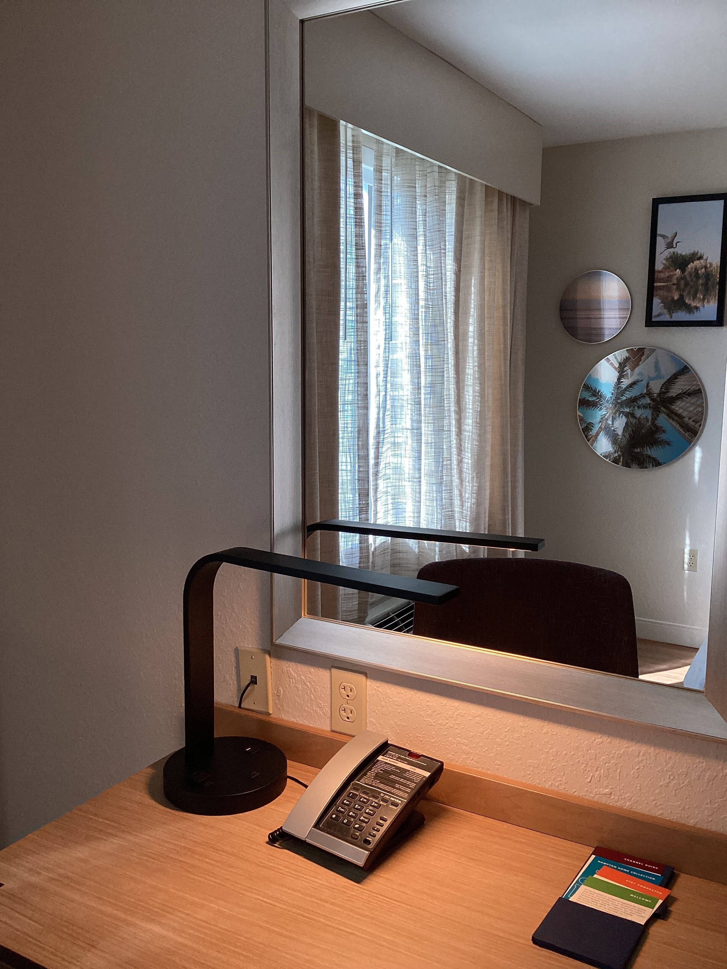 Een hotelkamer met een bureau, telefoon, spiegel en raam met gordijnen.