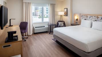 A hotel room with a large bed, a desk with a coffee maker, a chair, a TV, a window with curtains, and a view of buildings outside.