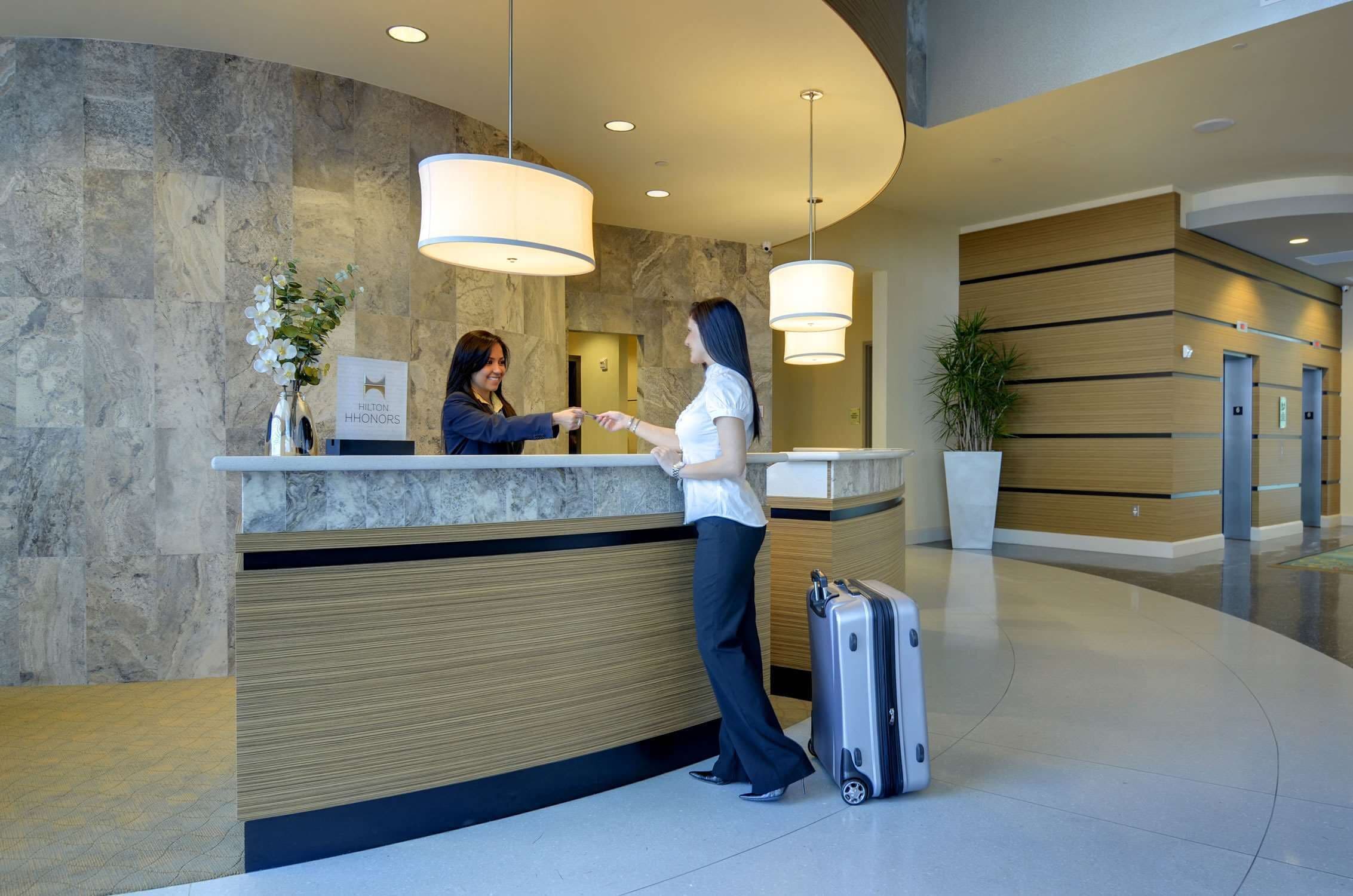 A hotel reception desk with a marble wall, wooden paneling, and a modern chandelier.