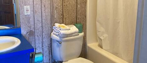 Un baño con pared de madera, inodoro blanco, lavabo azul y cortina de ducha.