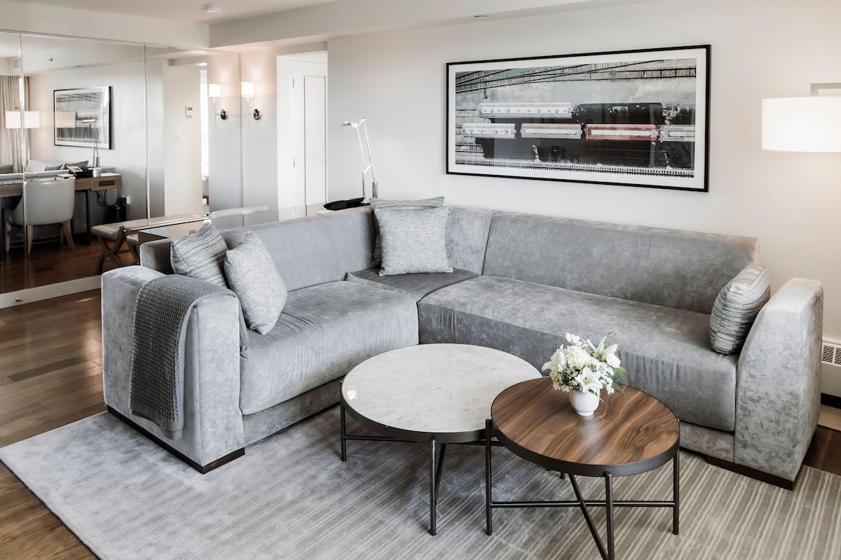 A modern living room with a grey sectional sofa, two round coffee tables, a framed artwork on the wall, and a dining area in the background.