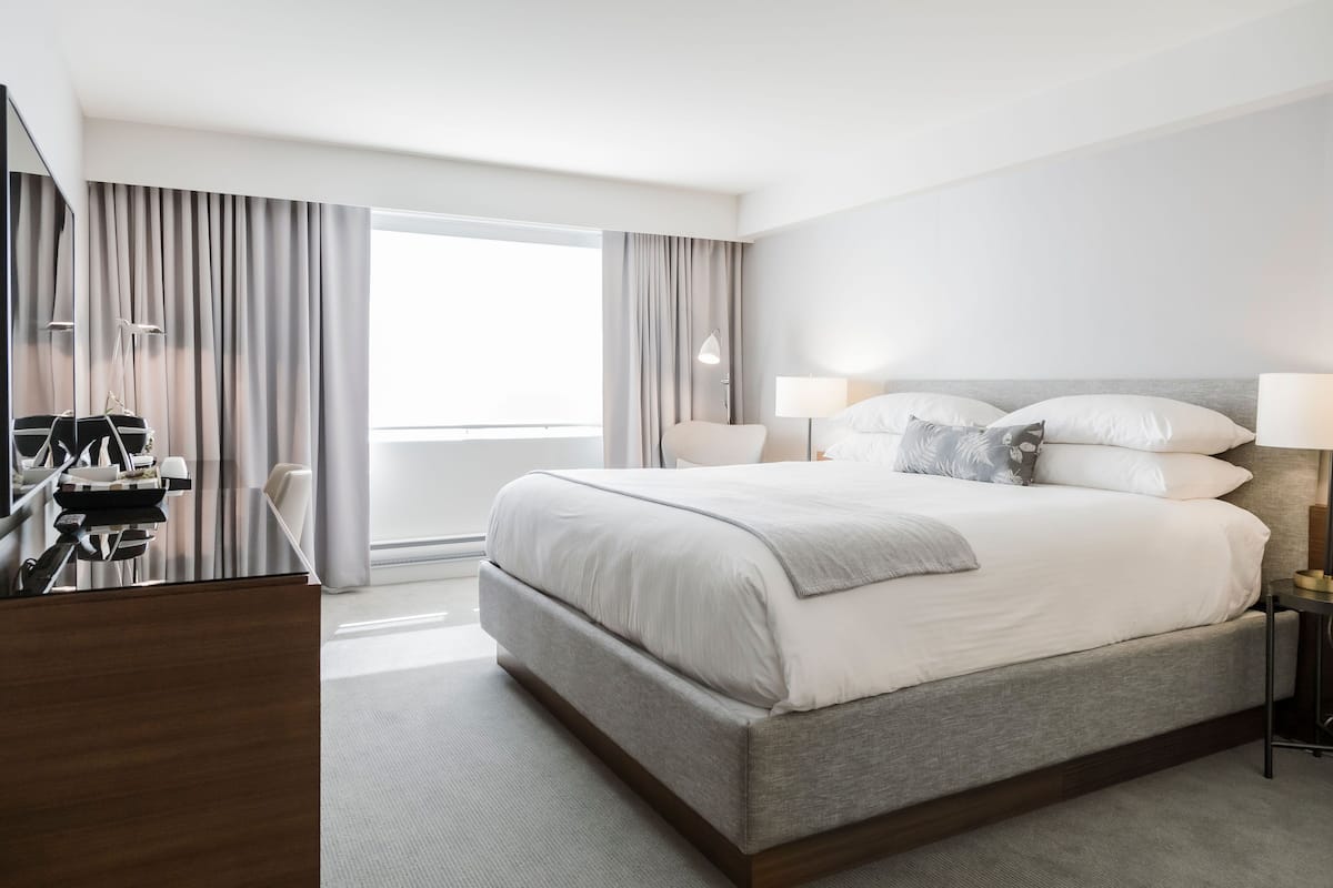 A modern hotel room with a large bed, a desk with a chair, and a large window.