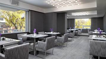 A modern restaurant with grey upholstered chairs and tables, large windows with a view of trees, and a ceiling with multiple light fixtures.