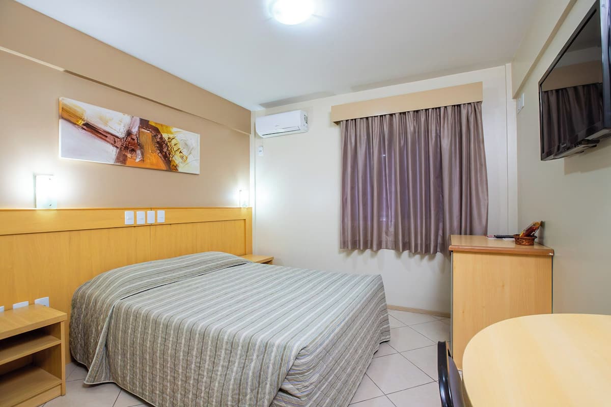 Hotelkamer met een bed, bureau, stoel, airconditioning, televisie en raam met gordijnen.