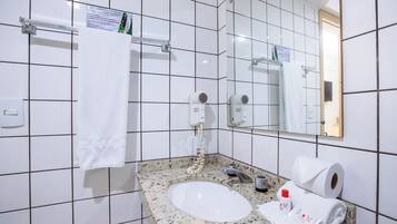 Un bany amb parets revestides de rajoles blanques, un lavabo amb taulell de granit, una tovallola penjada en un suport i un mirall, a més d'un dispensador de tovalloles.