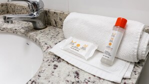 Une salle de bain avec un comptoir en granit, un lavabo blanc, un robinet argenté et une serviette blanche. On y trouve deux articles de toilette : un flacon blanc avec un bouchon orange et un sachet blanc avec du texte.