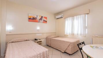 A room with two beds, a desk, a chair, a table, an air conditioning unit, a window with curtains, and a painting on the wall.