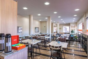 A spacious dining area with multiple tables and chairs, a coffee station with branded thermos cups, and a colorful box of snacks. - Hotel Express Canoas (Canoas)
