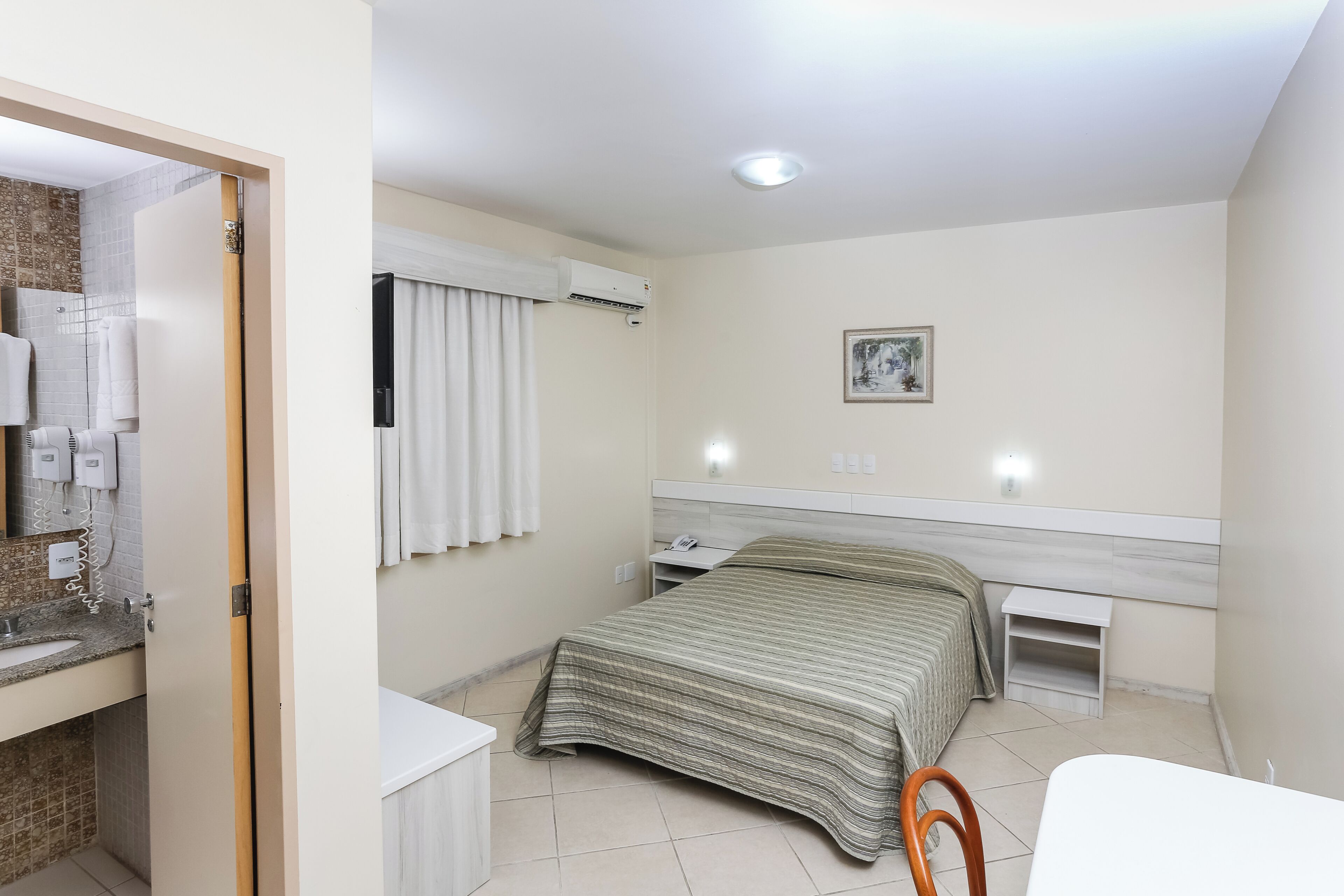 A bedroom with a bed, a bedside table, a nightstand, a small table, a chair, and a bathroom with a sink and towel rack.