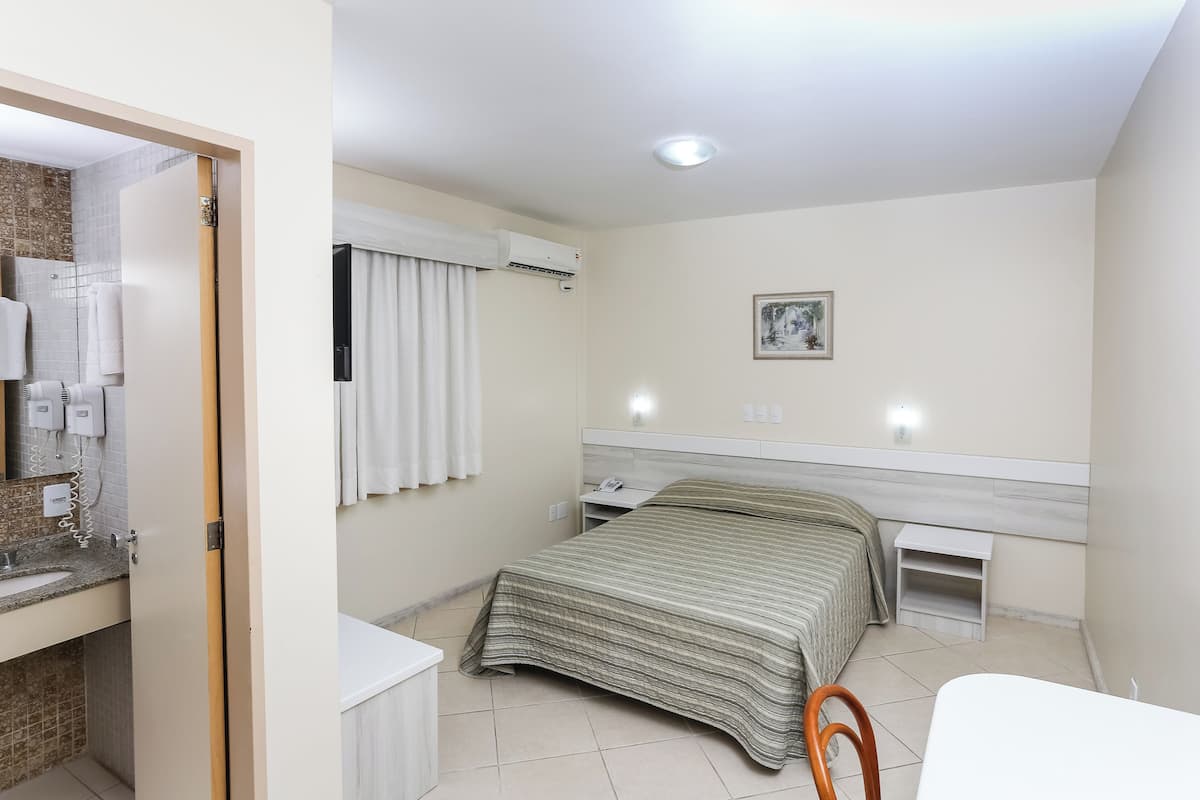 A bedroom with a bed, a bedside table, a nightstand, a small table, a chair, and a bathroom with a sink and towel rack.