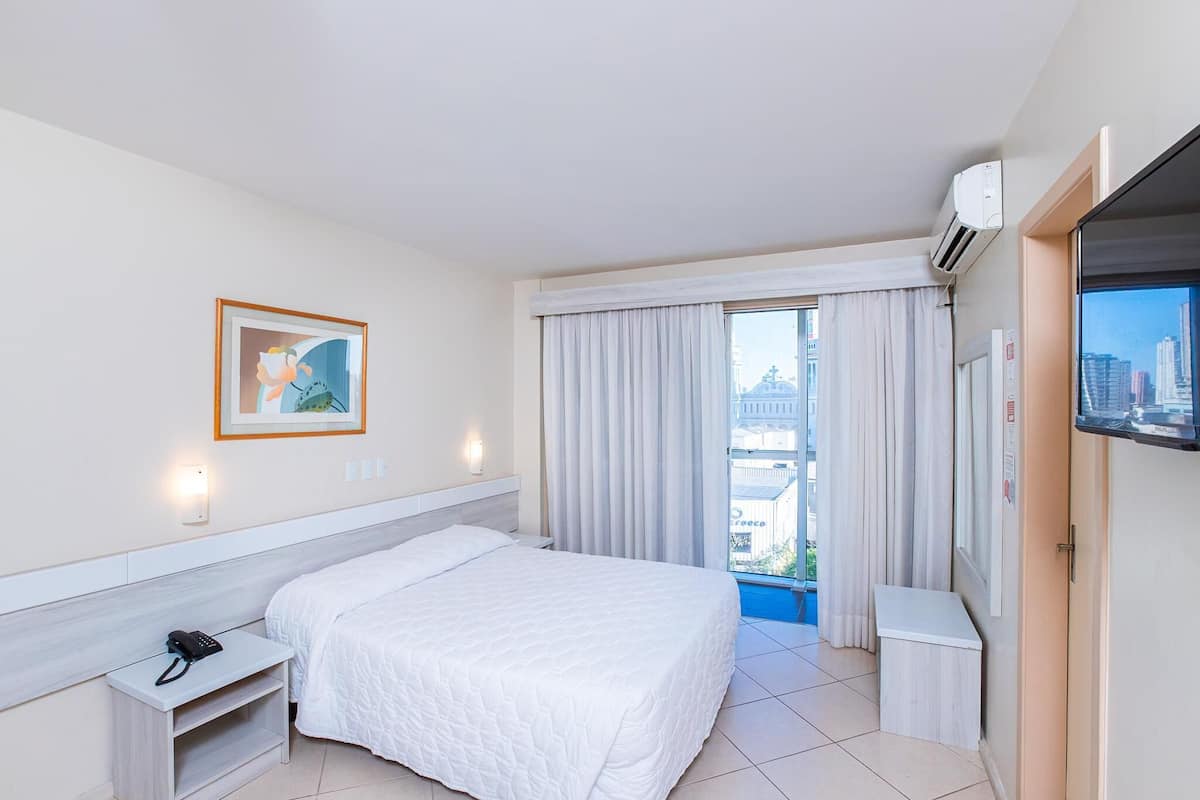 A hotel room with a bed, a nightstand, a TV, a window with curtains, and a city view.