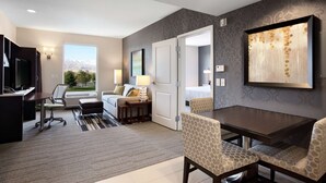 A hotel room with a dining area, a desk with a computer, a bed, and a large window with a mountain view. - Home2 Suites by Hilton Salt Lake City/West Valley City, UT (West Valley City)