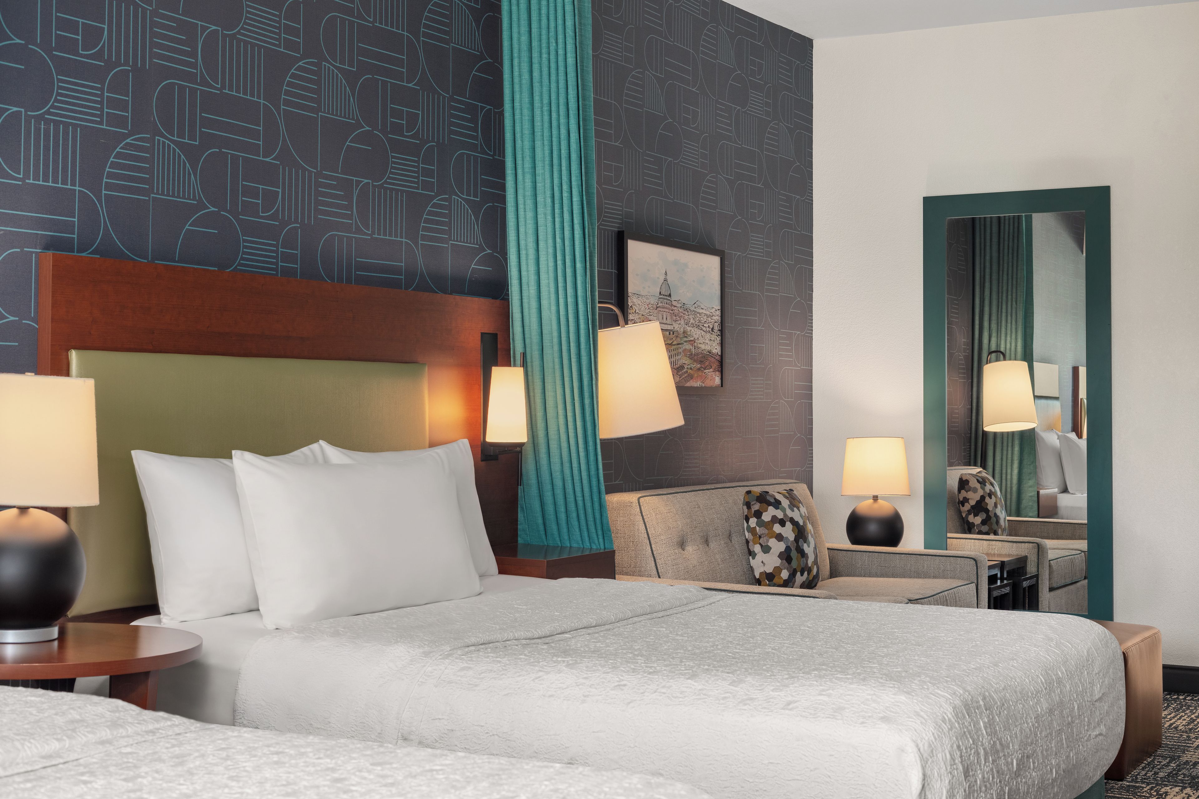 Ein Hotelzimmer mit einem großen Bett, zwei Nachttischen mit Lampen, einer Sitzecke mit einem Sessel, einem Spiegel und einer Wand mit geometrischem Muster.
