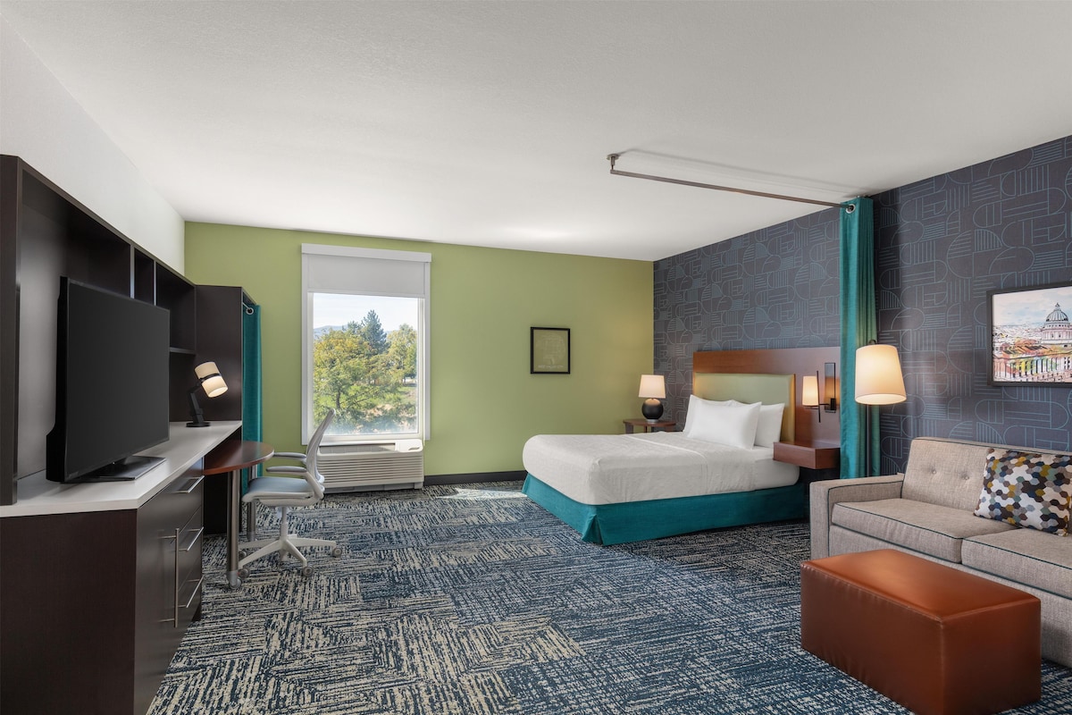 A hotel room with a bed, a desk with a TV, a chair, a sofa, and a window with a view of trees.