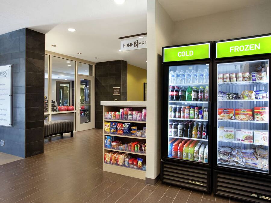 Un hall d’hôtel doté d’un espace de libre-service proposant des boissons froides et glacées, d’un coin salon et d’un panneau indiquant l’emplacement du comptoir d’enregistrement pour les clients séjournant à l’hôtel ou ceux qui s’y rendent pour affaires.