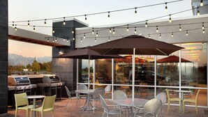 An outdoor patio with tables and chairs, string lights, and a grill area. - Home2 Suites by Hilton Salt Lake City/West Valley City, UT (West Valley City)