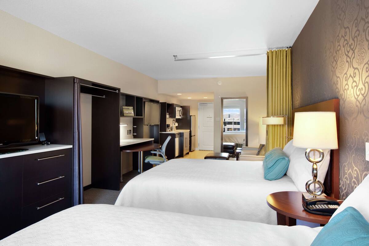A hotel room with two beds, a desk, a TV, and a kitchenette.