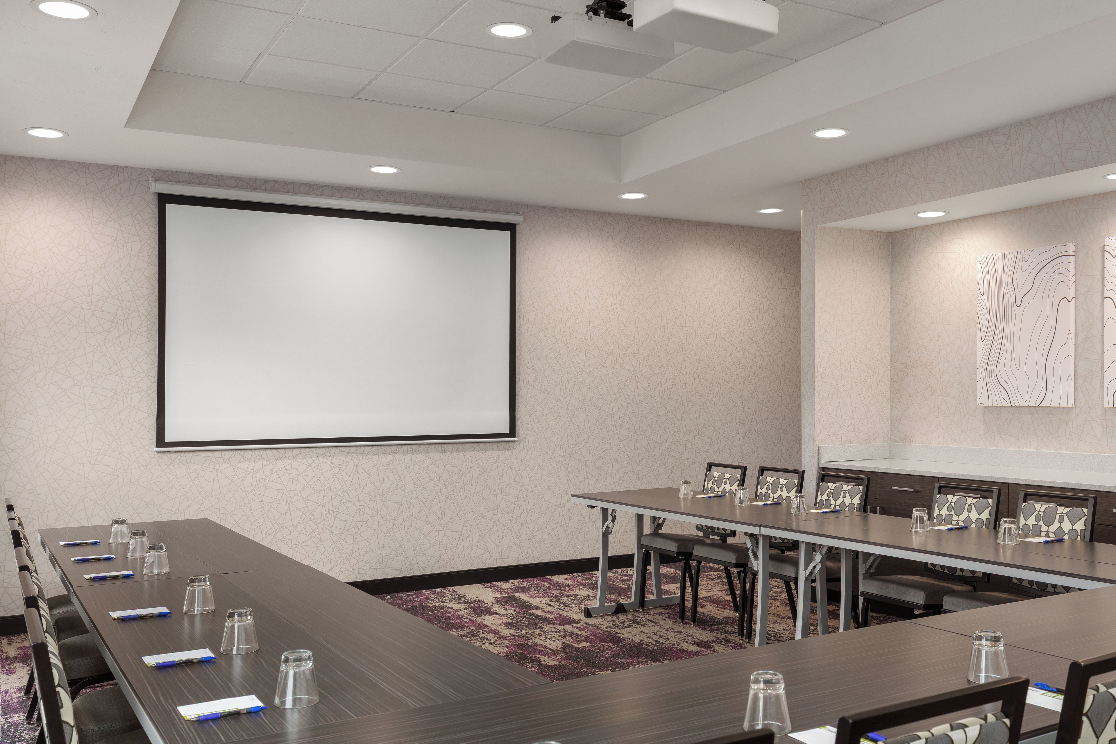 A conference room with a long table, chairs, a projector screen, and a carpeted floor.