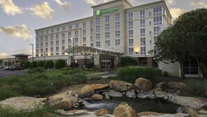 A Holiday Inn hotel with a landscaped entrance, a small stream, and a clear sky.