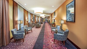 A hotel corridor with patterned carpet, seating areas, and wall-mounted lamps.