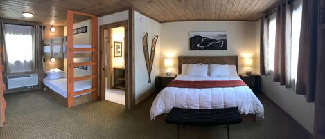 A hotel room with bunk beds, a large bed with a red blanket, a headboard, bedside lamps, a wall-mounted picture, and a wooden ceiling.