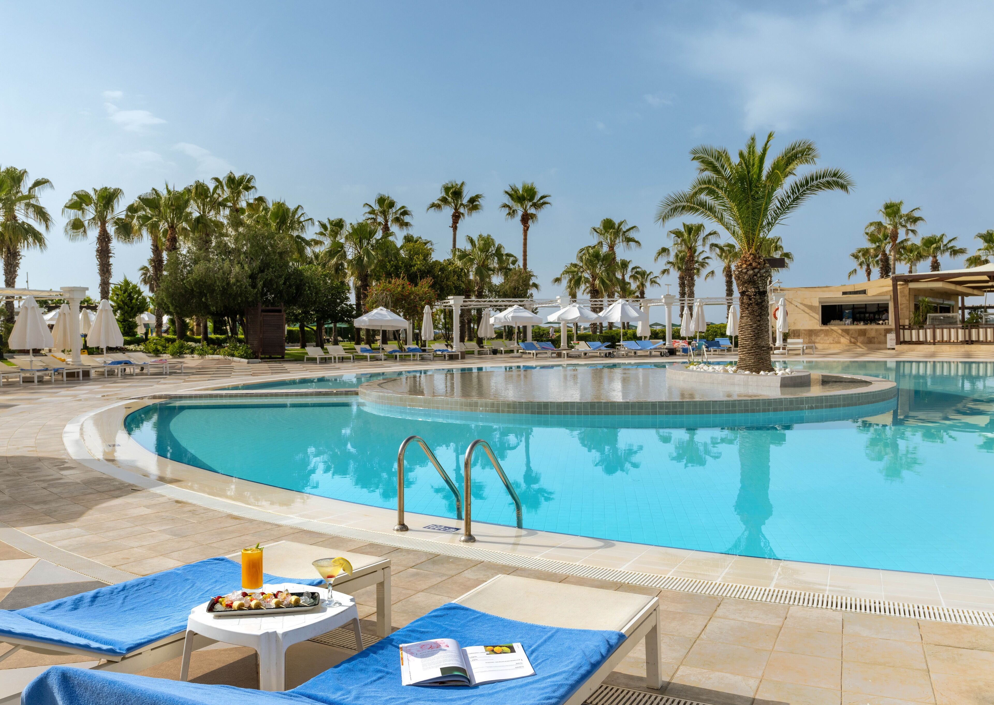 Zona junto a la piscina con sillones de descanso, sombrillas y una mesa de comedor con comida y bebidas.