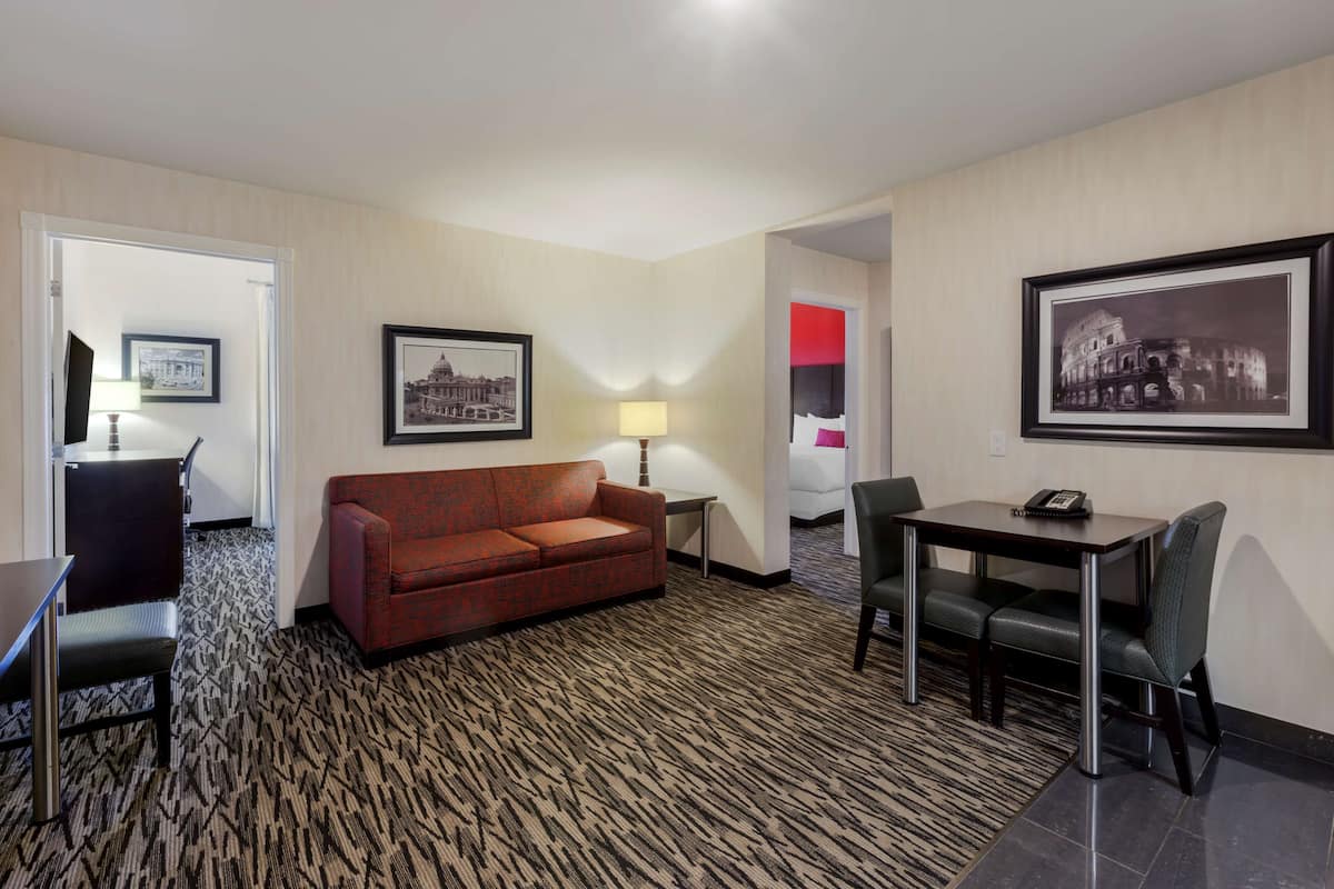 Habitación de hotel con alfombra estampada, sofá rojo, mesa de comedor con sillas y cuadros en las paredes.