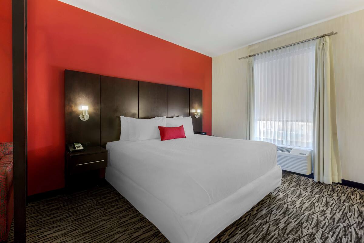 A hotel room with a large bed, a red accent wall, a dark headboard, bedside lamps, a telephone, and a window with curtains.