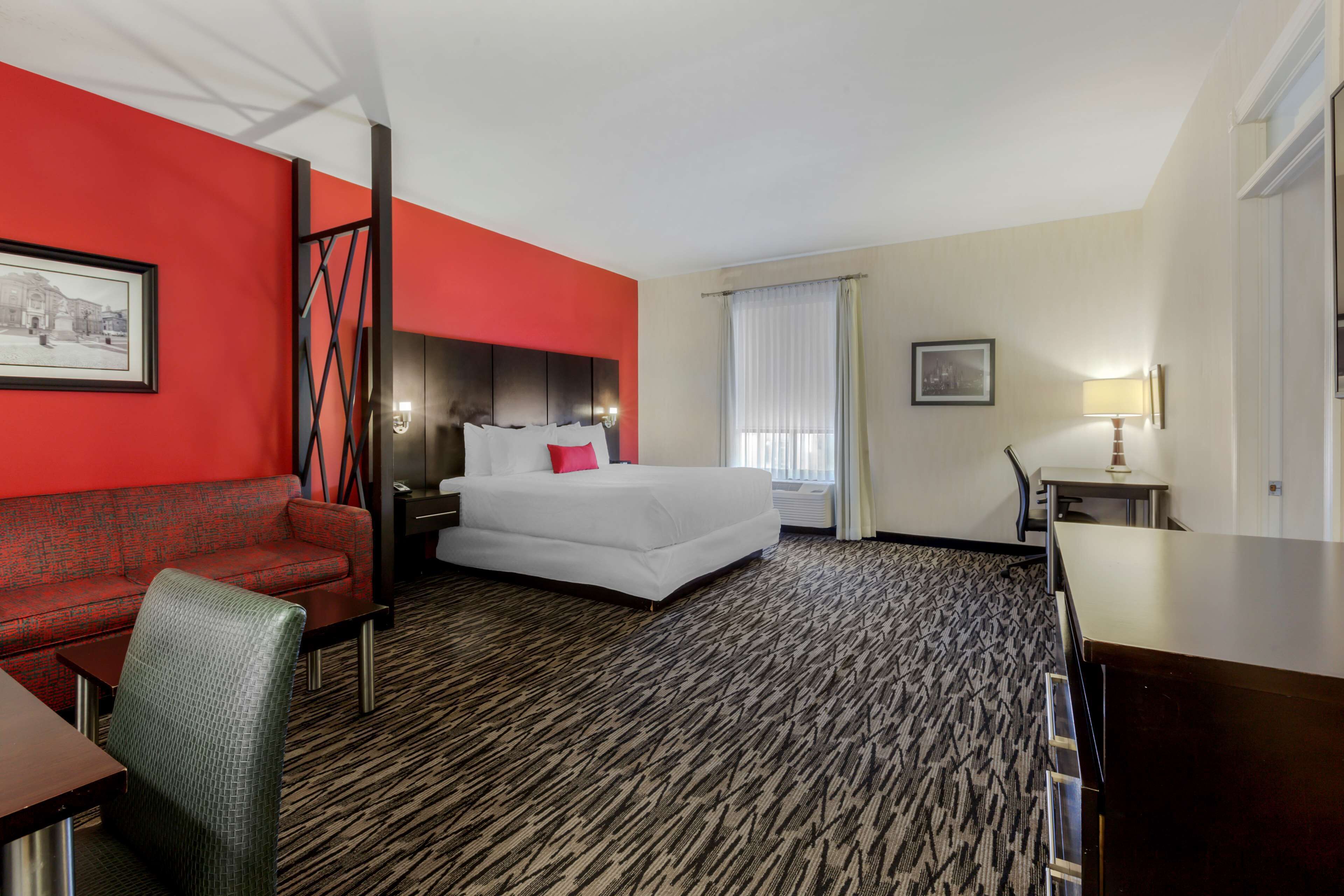 A hotel room with a red accent wall, a large bed, a desk, a chair, and a window with curtains.