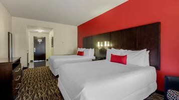 A hotel room with two beds, a red accent wall, a patterned carpet, and a desk with drawers.