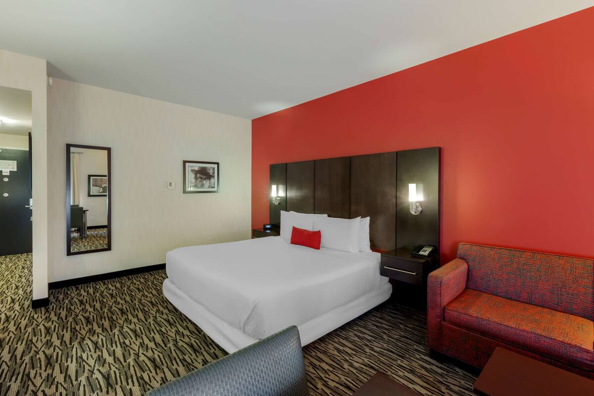 A hotel room with a large bed, a red accent wall, a seating area with a red and orange patterned sofa, and a patterned carpet.